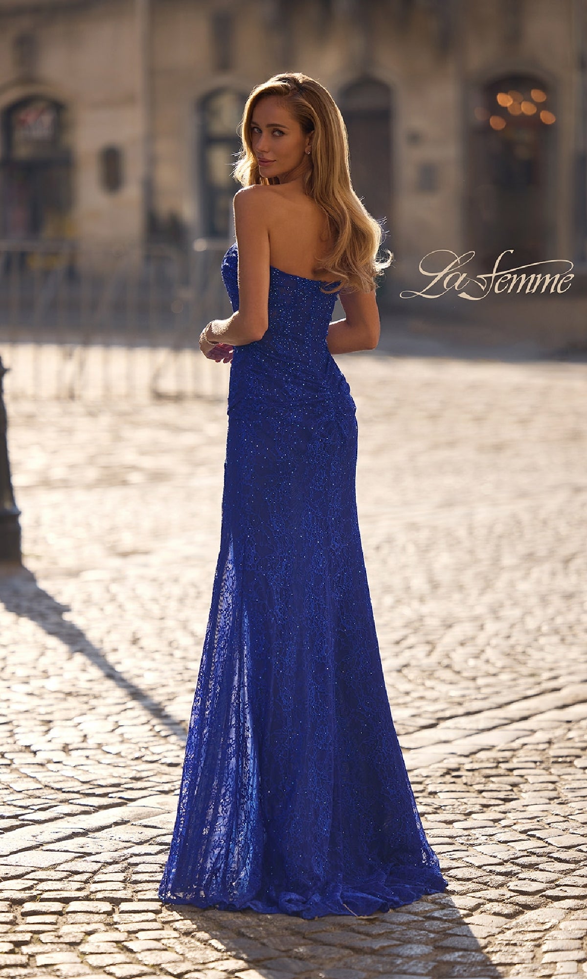 A woman stands outdoors on a cobblestone street, looking over her shoulder in the elegant Strapless Long Beaded Prom Dress: La Femme 32773, with